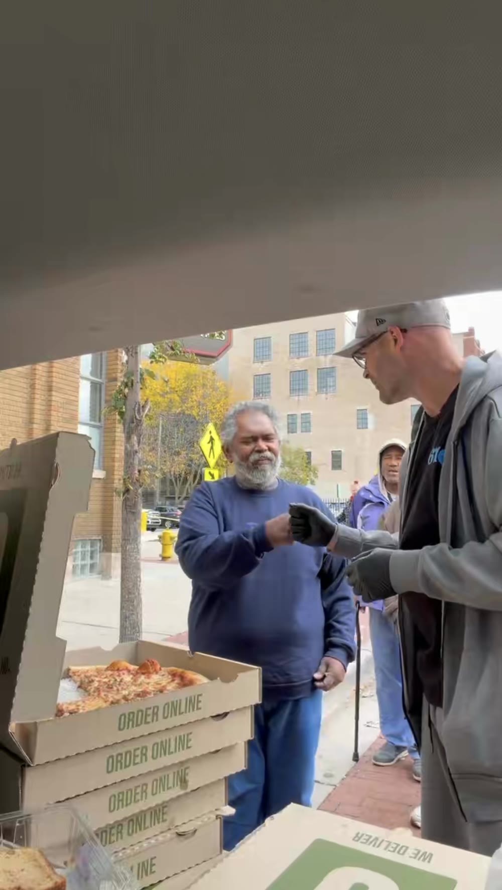 Tom hands a boxed meal to a man on a sunny sidewalk during a street outreach